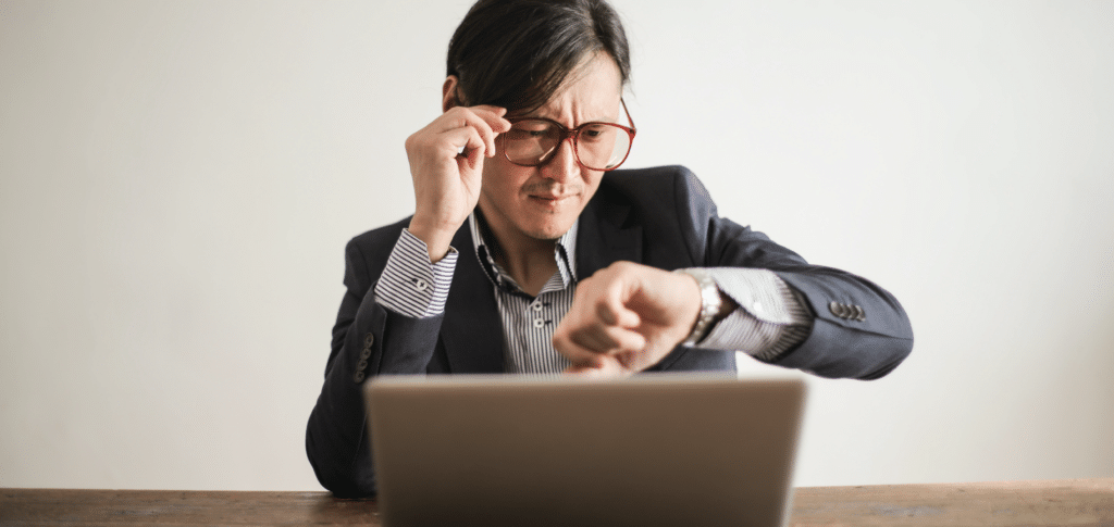 man straining eyes, lifting glasses while squinting at watch & laptop in front of him