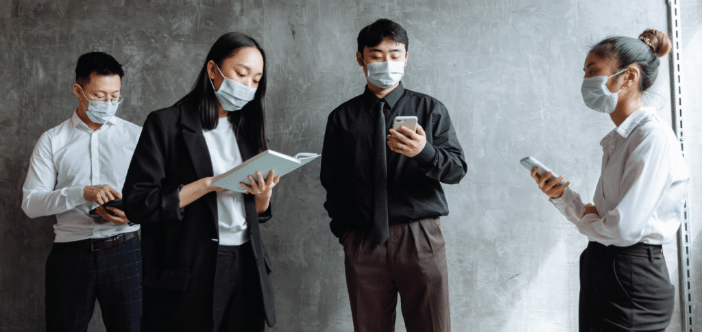 four people dressed in business casual wear & face masks standing and looking at tech devices