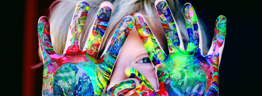 child with finger paint on hands in front of their face with eye peeking through