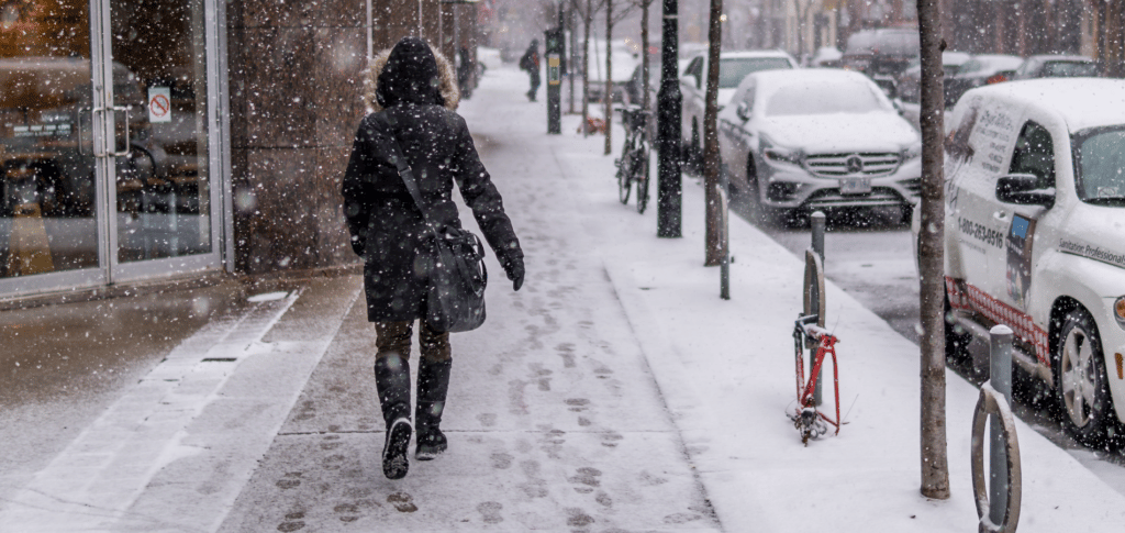 Person wearing black winter coat walking away from camera on snowy sidewalk